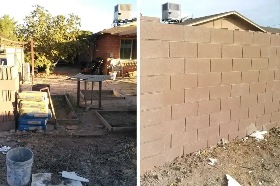 Partly built cinder block wall beside a dirt yard with construction materials and a house in the background
