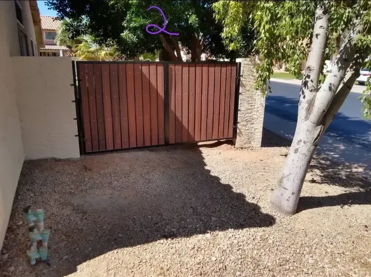 Closed brown wooden gate beside a gravel yard and tree, with a driveway and sidewalk beyond
