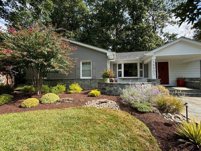 A single-story house with grey siding, a white porch, and a red door, surrounded by lush landscaping and a manicured lawn.