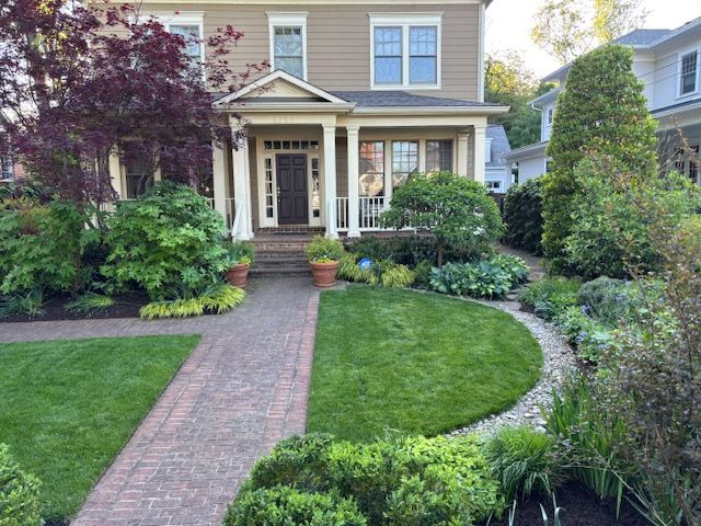 A two-story tan house with a brick walkway, lush green lawn, and manicured landscaping under a bright sky.