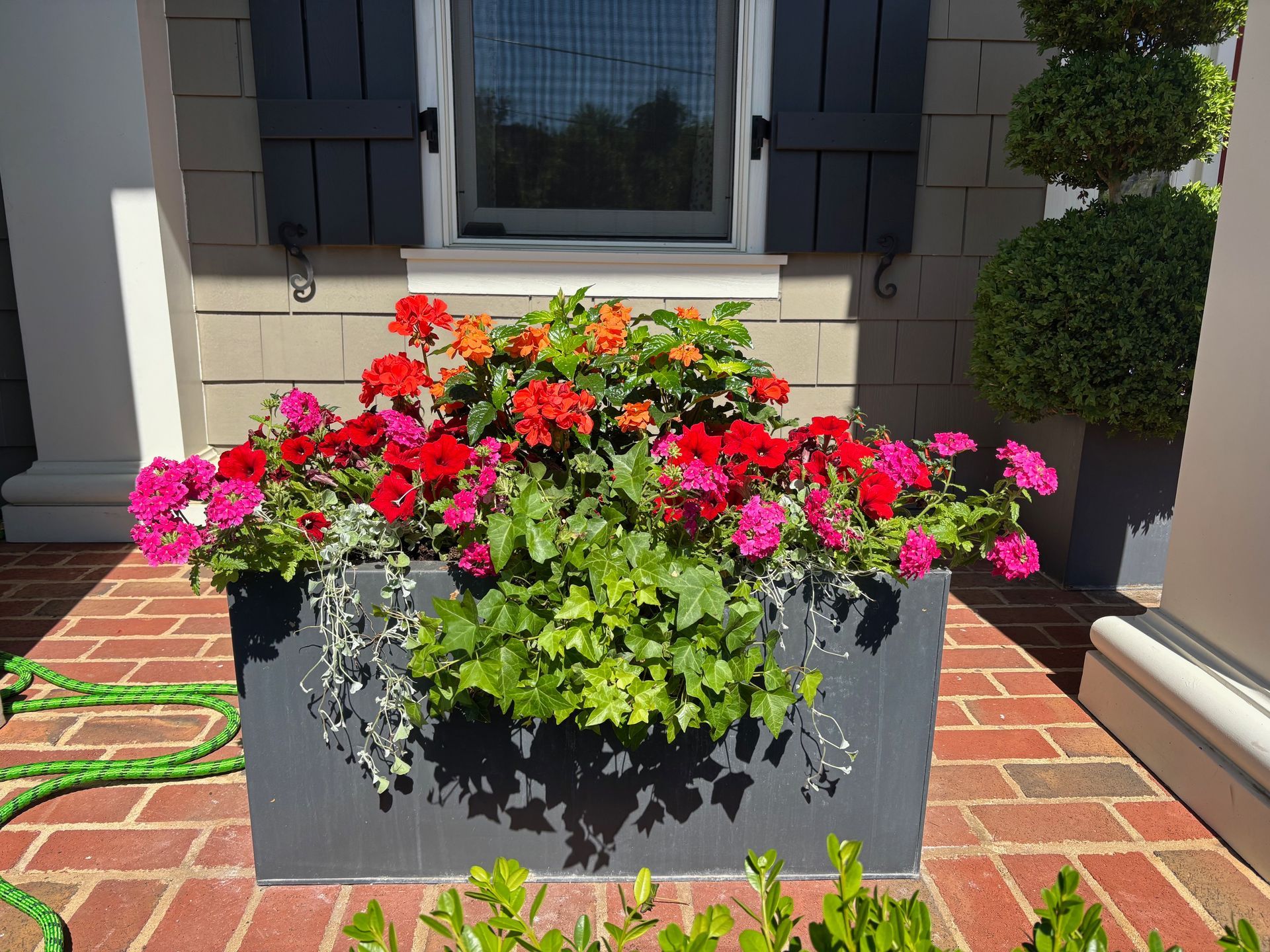 A rectangular grey planter box filled with vibrant red, pink, and orange flowers on a brick patio.