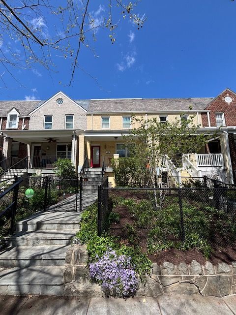 A street view of a row of brick and stone townhomes with front porches, stairs, and small gardens on a sunny day.