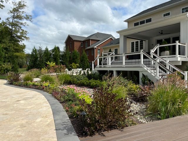 A backyard landscape featuring a stone patio, wooden deck, stairs, lush garden beds, and two houses on a cloudy day.