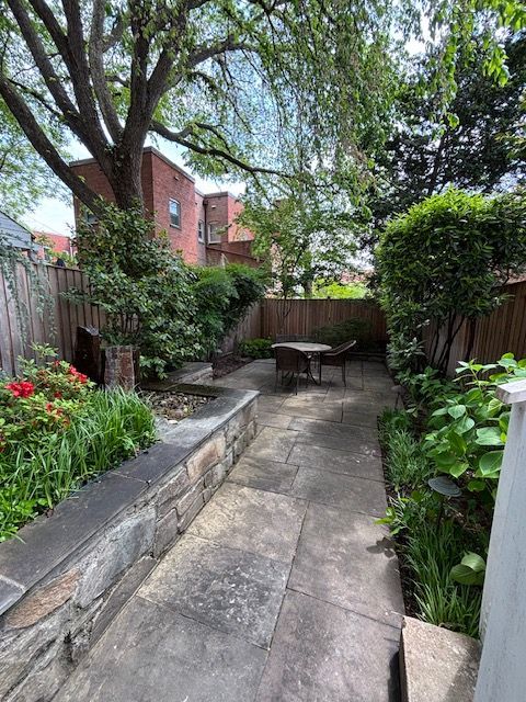 A paved backyard patio featuring a stone retaining wall, lush green plants, and a small dining set under a large tree.
