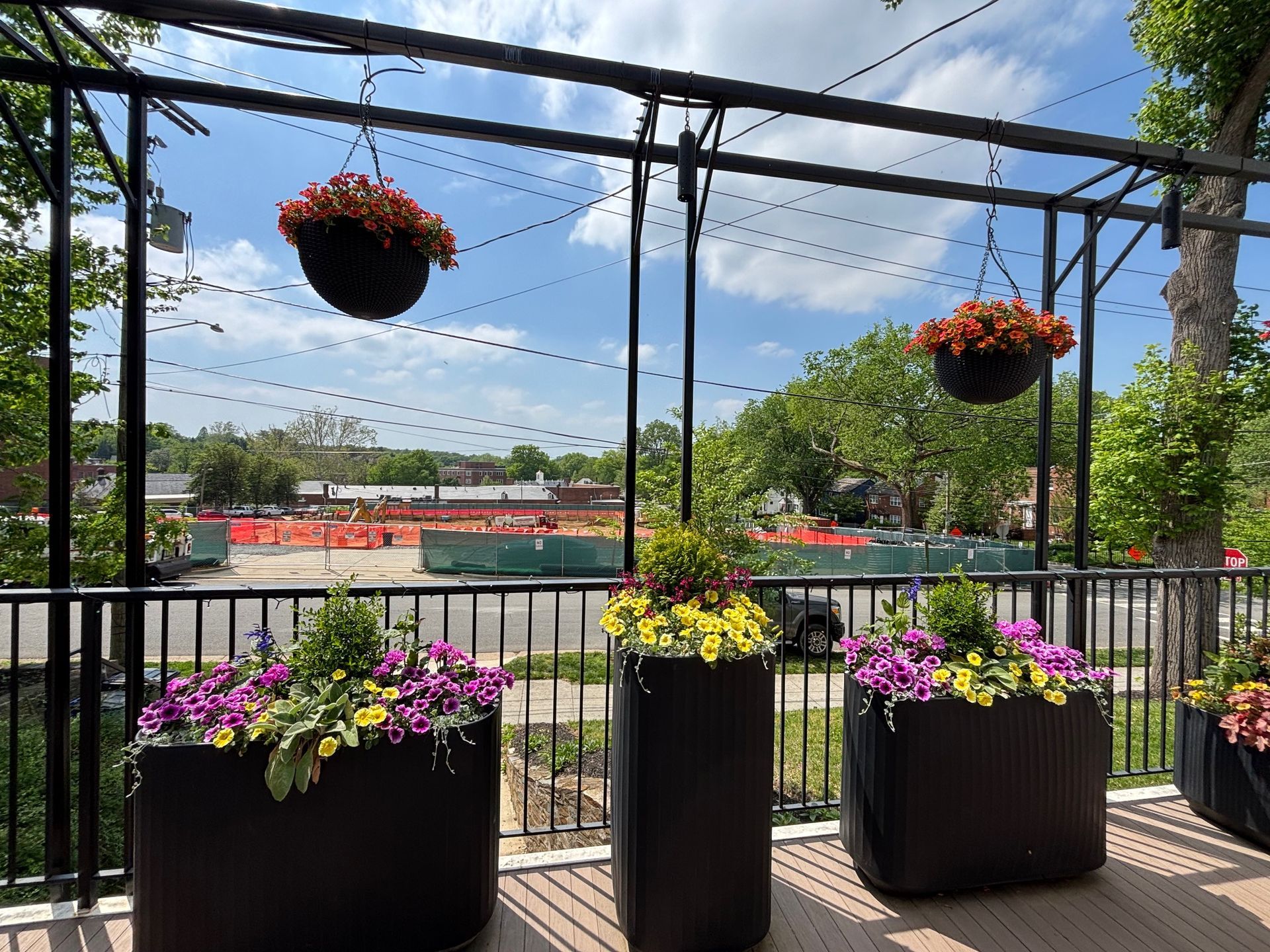 Black planters and hanging baskets filled with colorful flowers on a balcony overlooking a construction site.