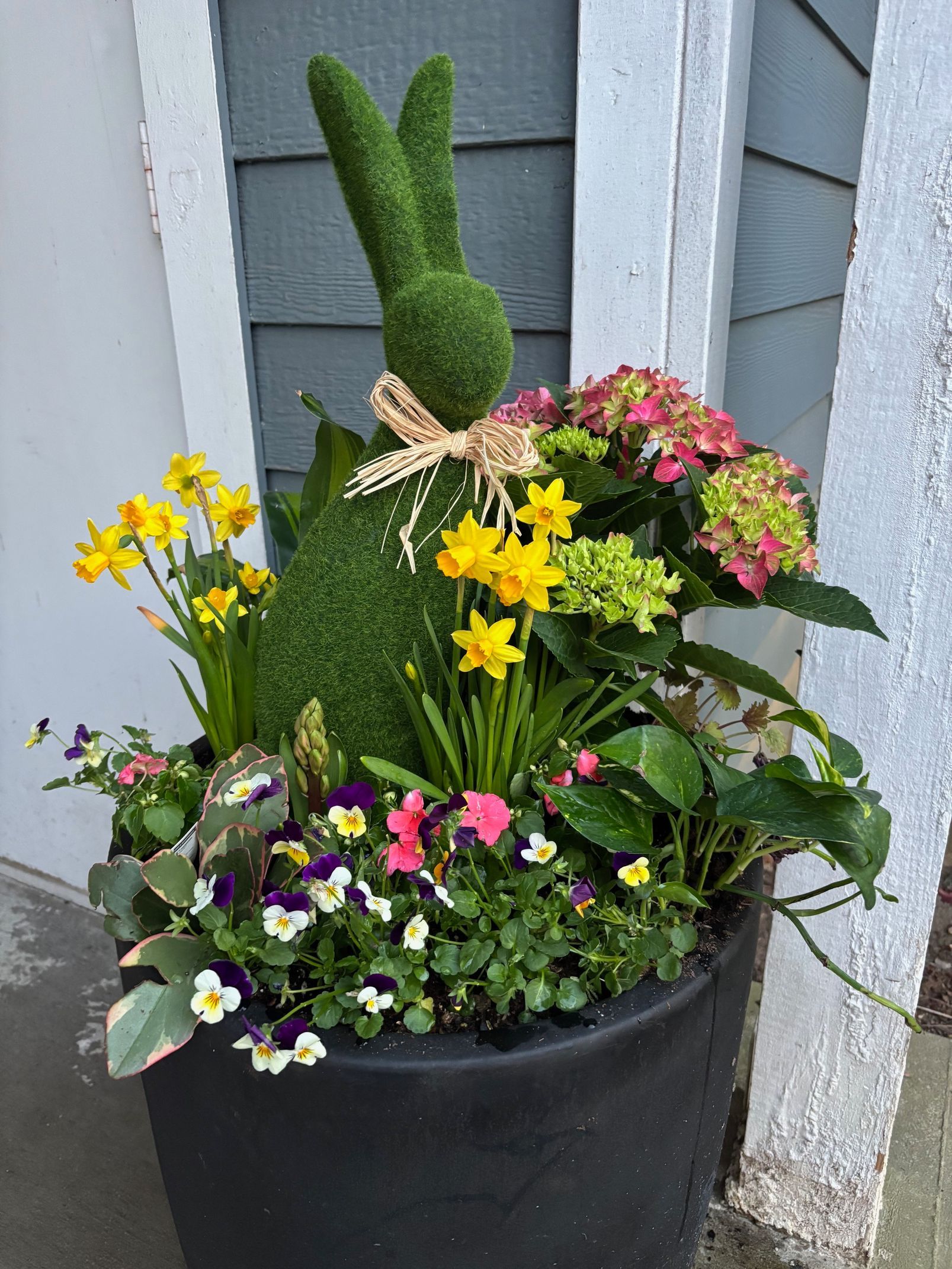 A black planter filled with a moss-covered rabbit, yellow daffodils, pink hydrangeas, and purple and white pansies.