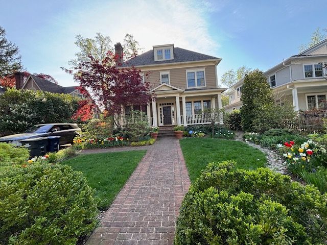 A two-story tan house with a brick path leading to the front door, surrounded by a green lawn and blooming gardens.