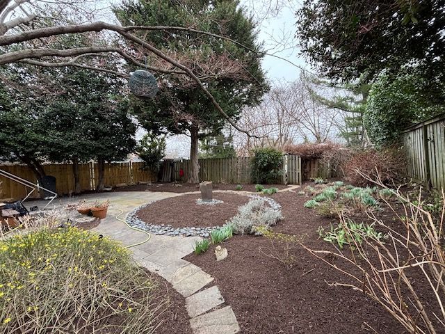 A residential backyard with a paved stone path leading to a central circular garden bed surrounded by mulch and trees.