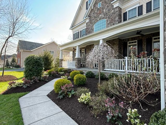 A concrete path leads to a large, multi-story stone and tan house with a covered front porch and landscaped garden beds.