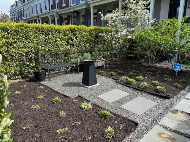 A small front garden with a gravel patio, two stepping stones, a black bench, a birdbath, and mulched planting beds.