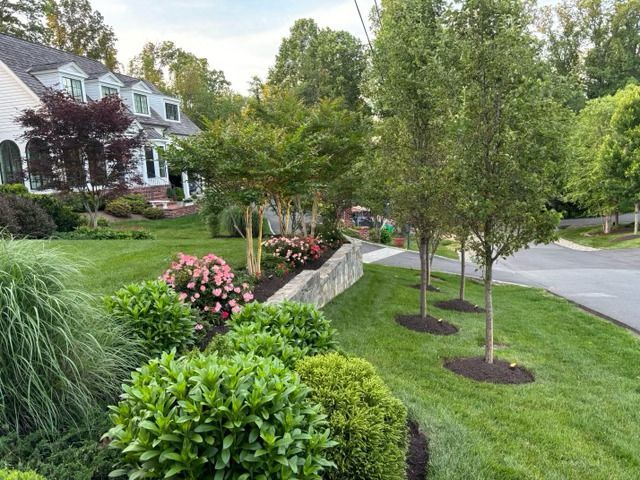 A landscaped front yard features a stone retaining wall, flowering bushes, and trees bordering a house and driveway.