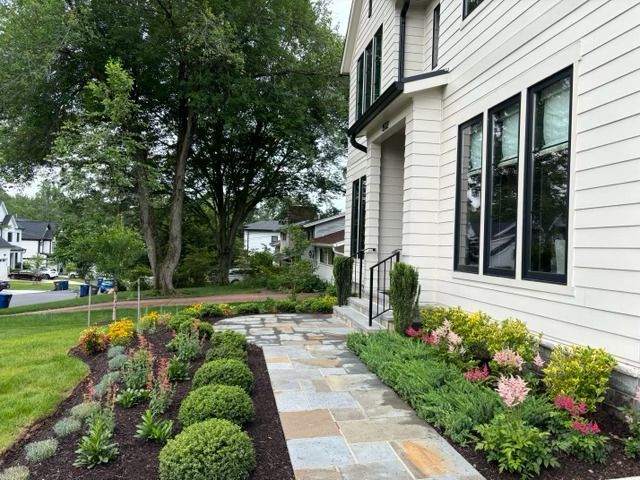 A stone walkway leads to the entrance of a modern white house with black window frames, flanked by landscaped gardens.