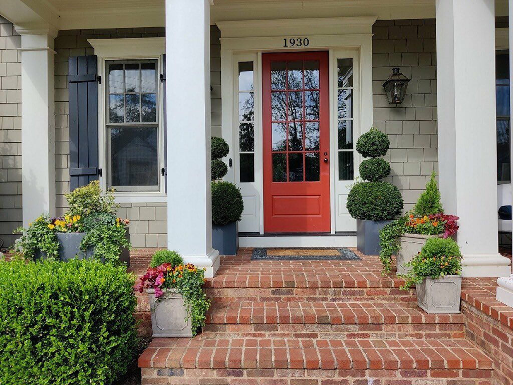 Front porch with brick steps, a bright red front door, and potted shrubs and flowers in a suburban setting.