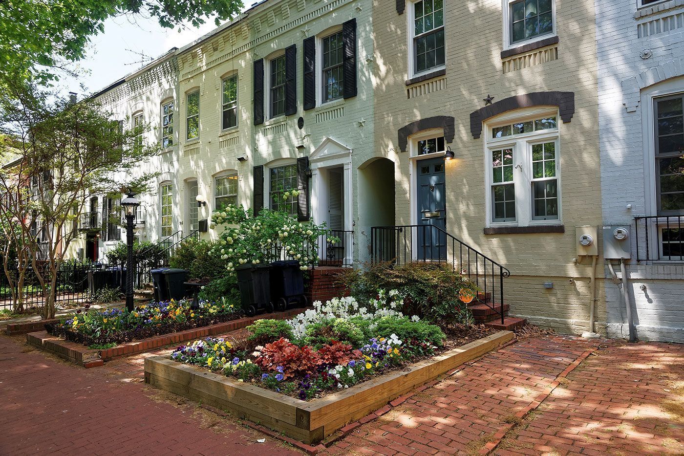A row of pale yellow and white historic brick townhouses with colorful flower beds and a red brick patio in front.