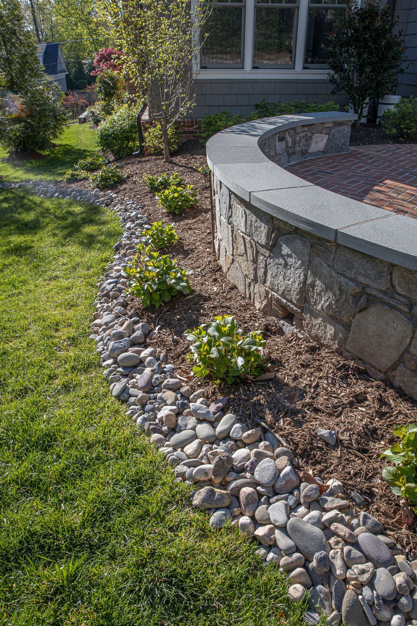 A curved stone retaining wall borders a garden bed with mulch, small green shrubs, and a decorative river rock edge.