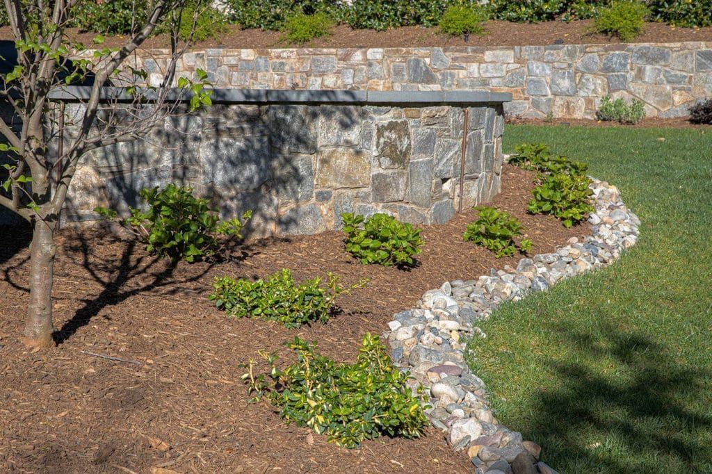 A landscaped garden with a curved stone retaining wall, small shrubs, and a mulch bed bordering a green lawn.