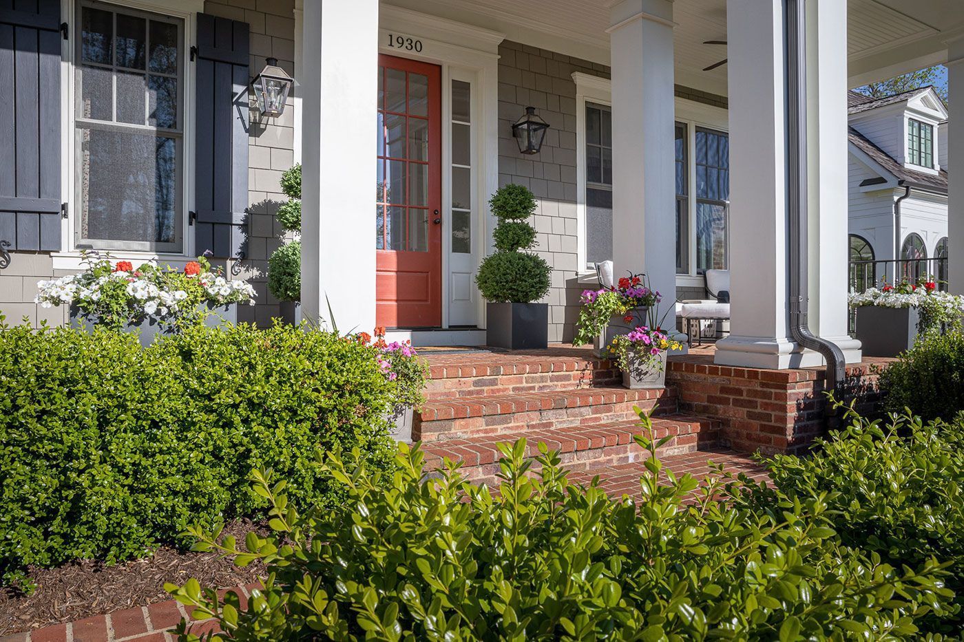 Front porch of a home with a red door, white columns, brick steps, and lush green bushes in the foreground.