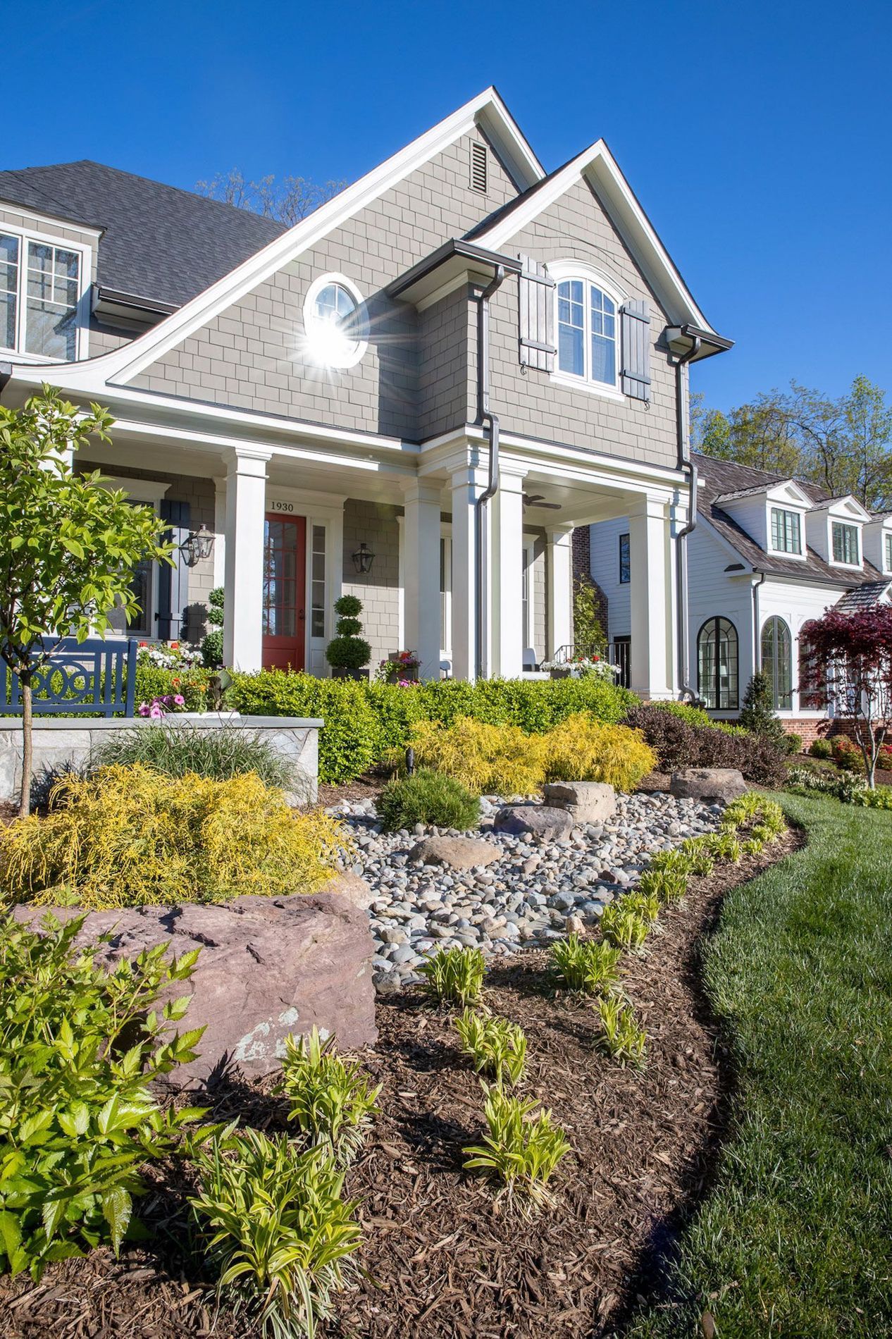 A gray house with a white porch and pillars features a landscaped front yard with mulch, rocks, and green shrubbery.