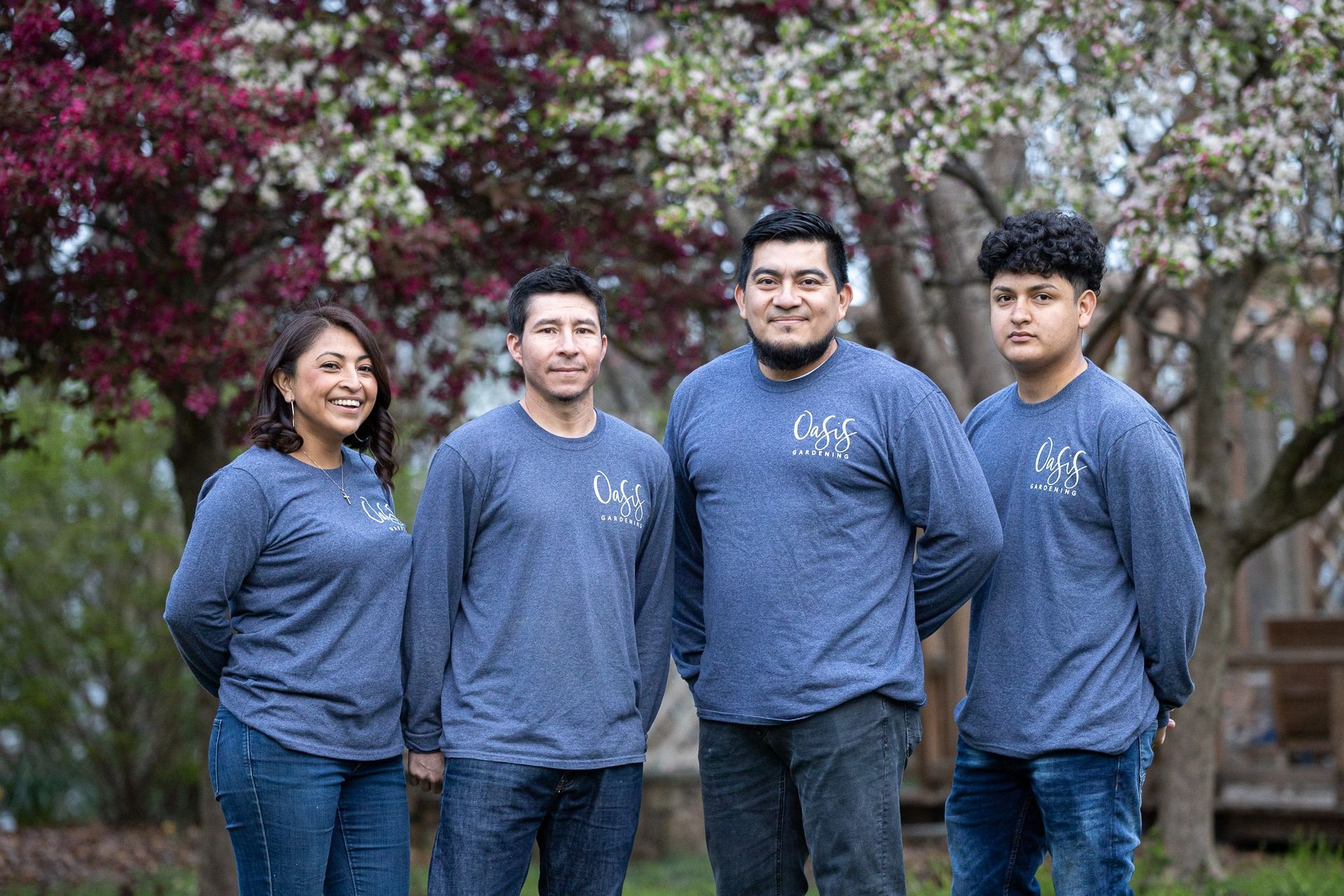Four people wearing matching blue long-sleeved shirts stand together outdoors in front of blooming trees.