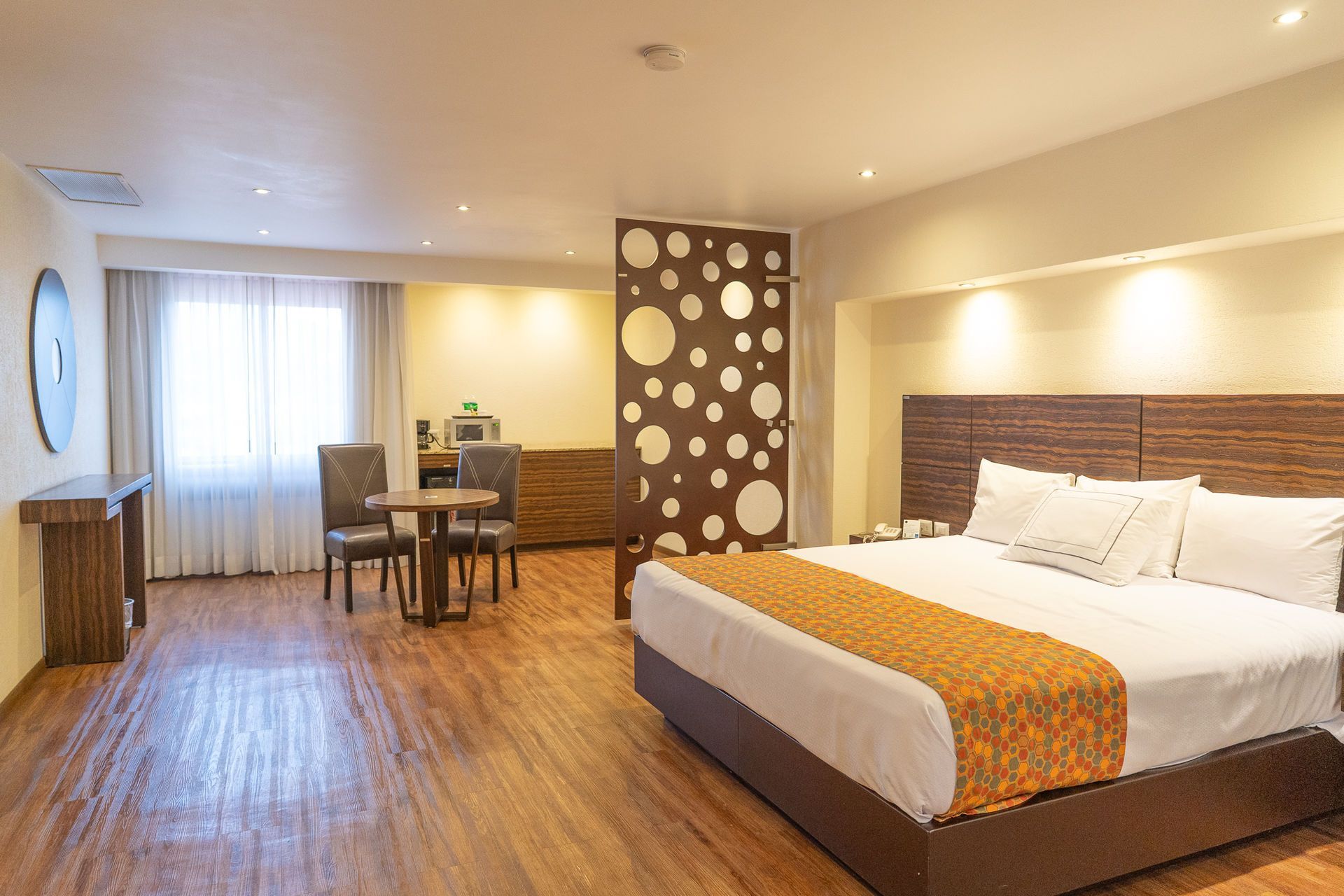 Una habitación de hotel con una cama grande, una mesa con dos sillas y un biombo decorativo con un diseño circular.