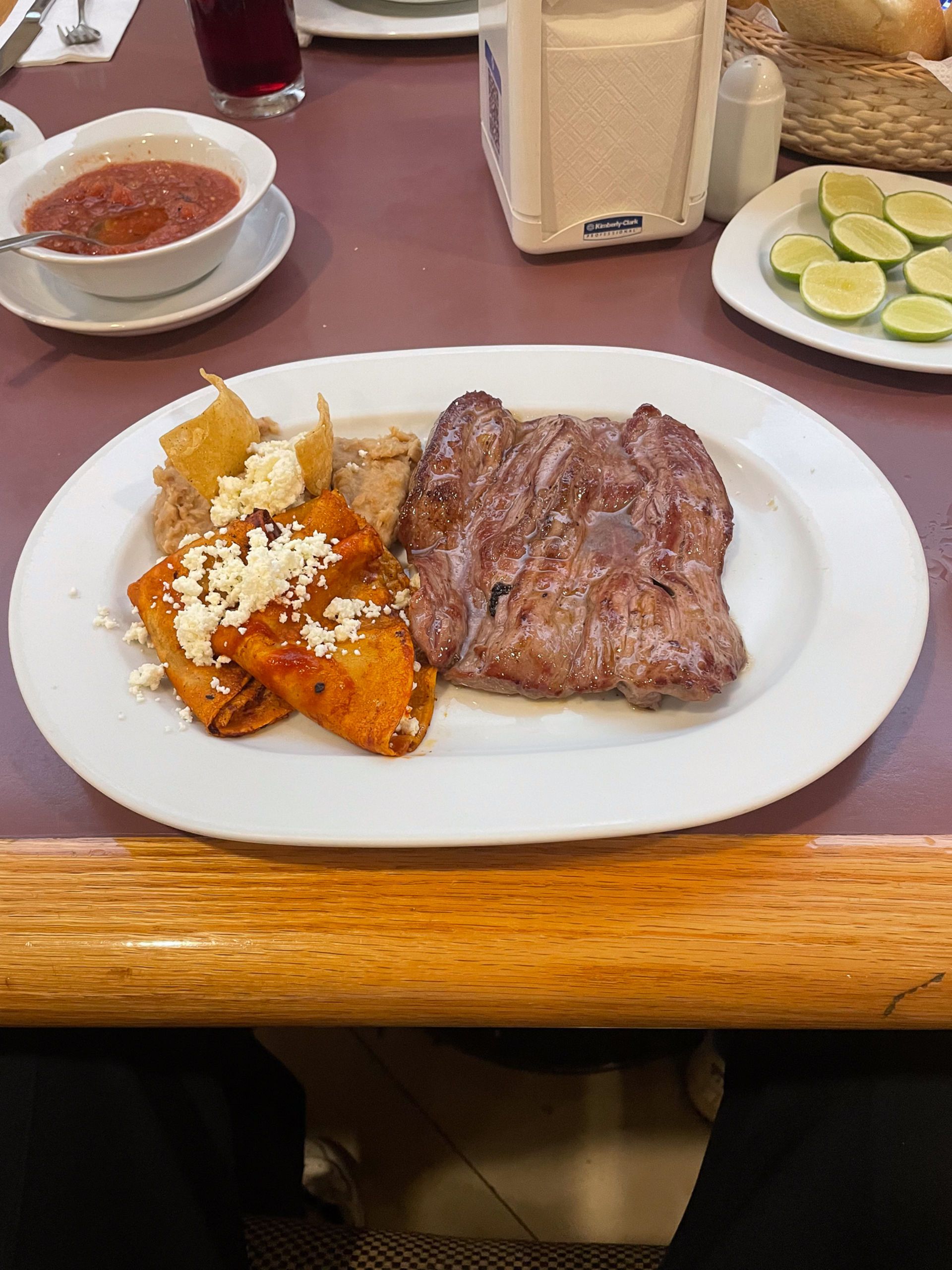 Un plato que incluye bistec a la parrilla, enchiladas rojas cubiertas con queso y frijoles refrito