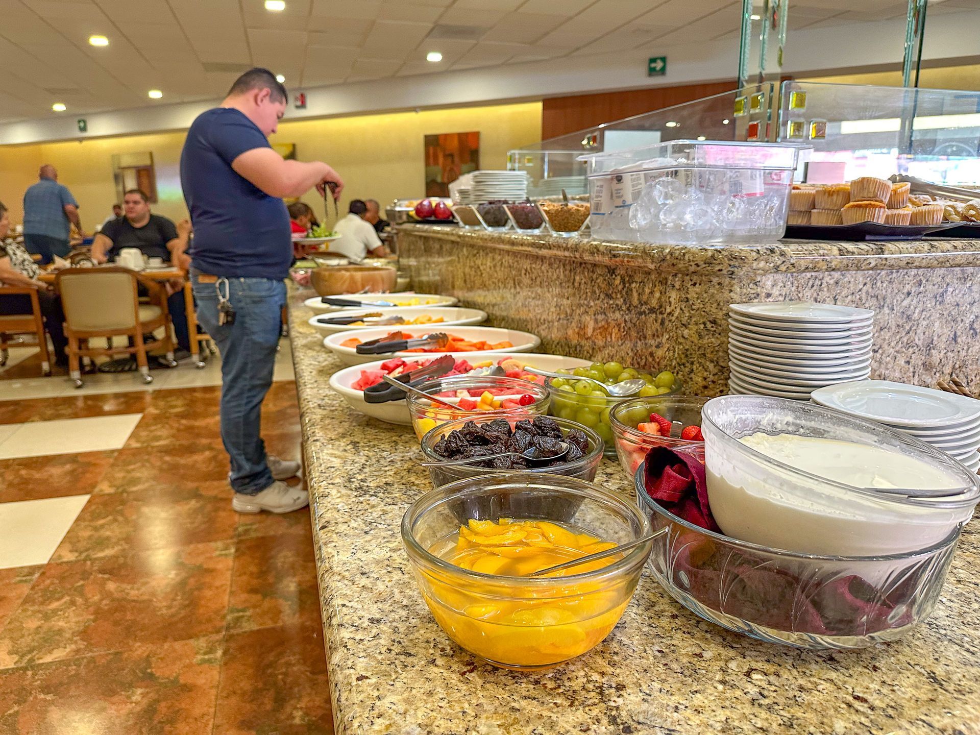 Un mostrador tipo bufé repleto de cuencos de fruta, yogur y platos de pan, con una persona de pie seleccionando la comida.