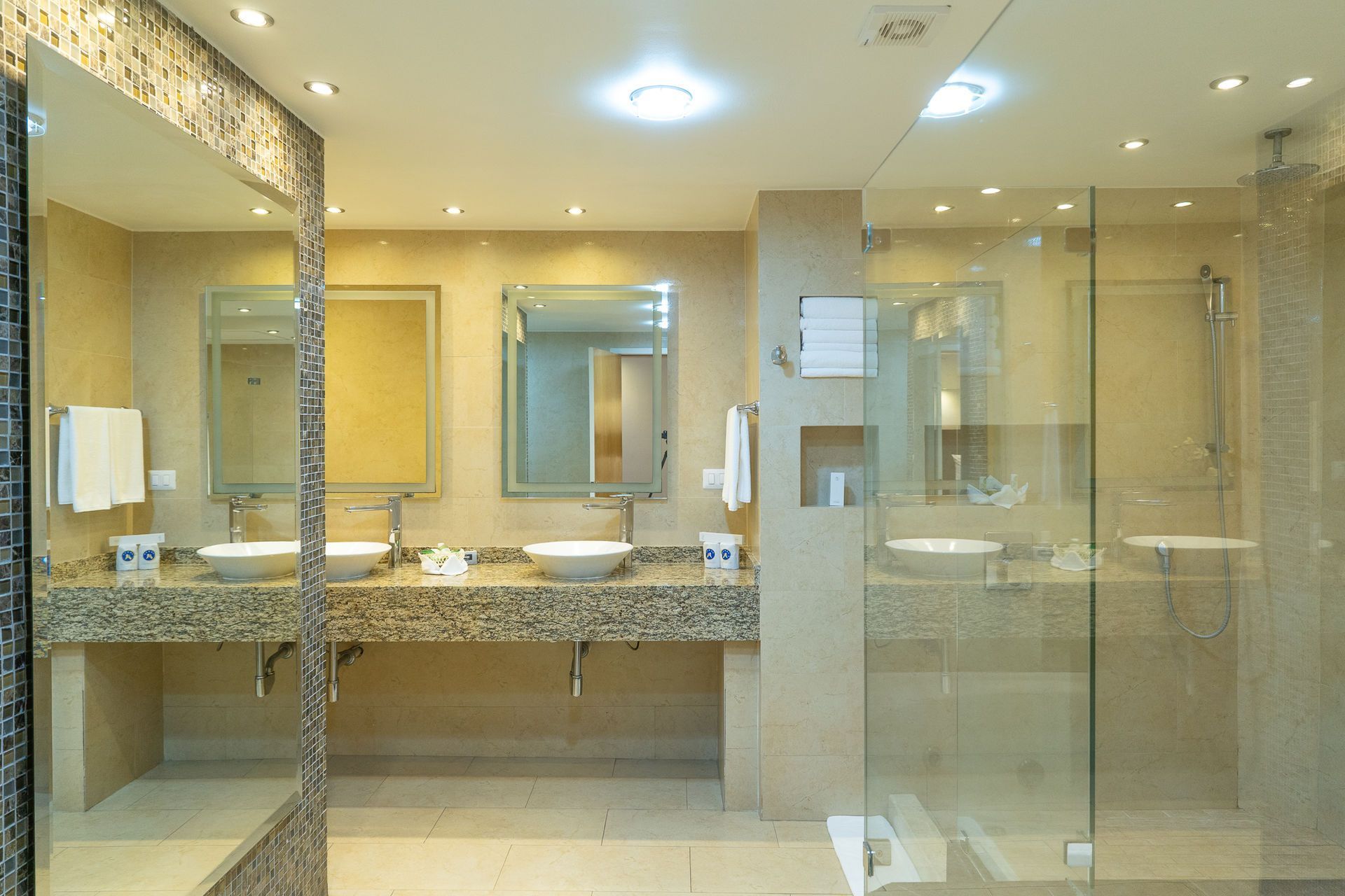 Un moderno baño de hotel con dos lavabos con encimeras de mármol beige, dos espejos y una ducha con mampara de cristal.