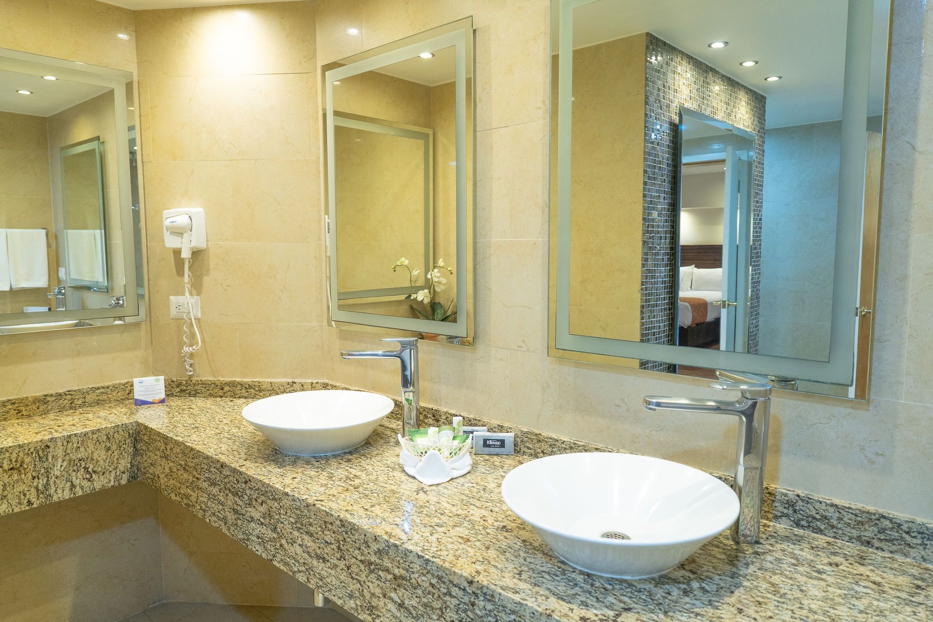 Un tocador de baño de hotel con dos lavabos blancos, grifos cromados y espejos montados sobre una encimera de granito beige.