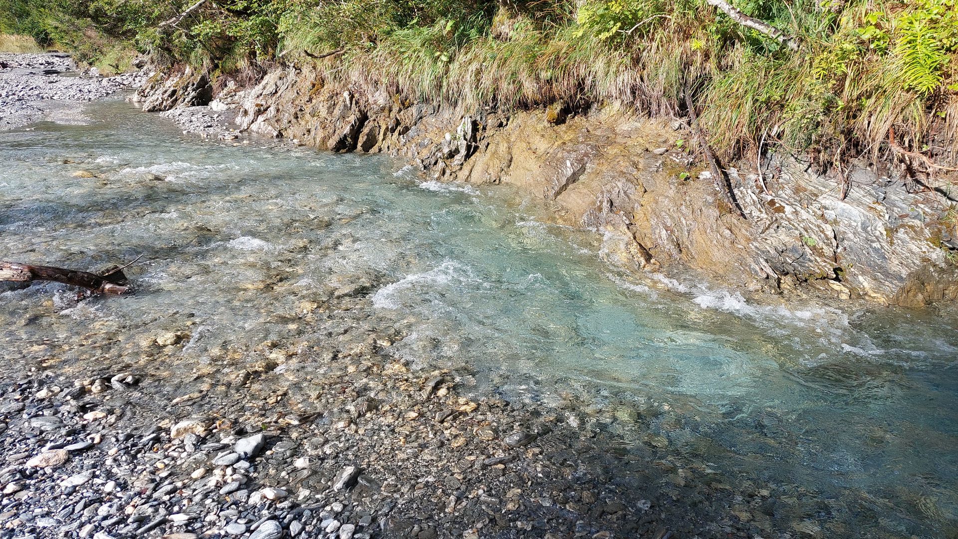 Klarer, fließender Bach mit steinigem Bett, der von einem Erdwall und grünem Gras gesäumt ist.