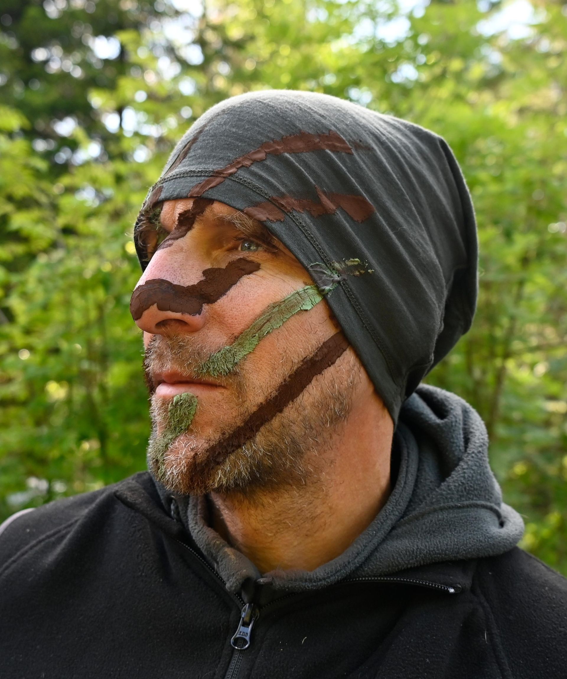 Mann mit Tarngesichtsbemalung und Hut in einer Waldlandschaft.