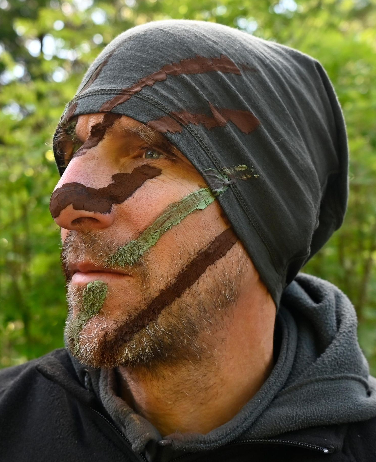 Ein Mann mit grauer Mütze und Tarngesichtsbemalung in einem Waldgebiet.