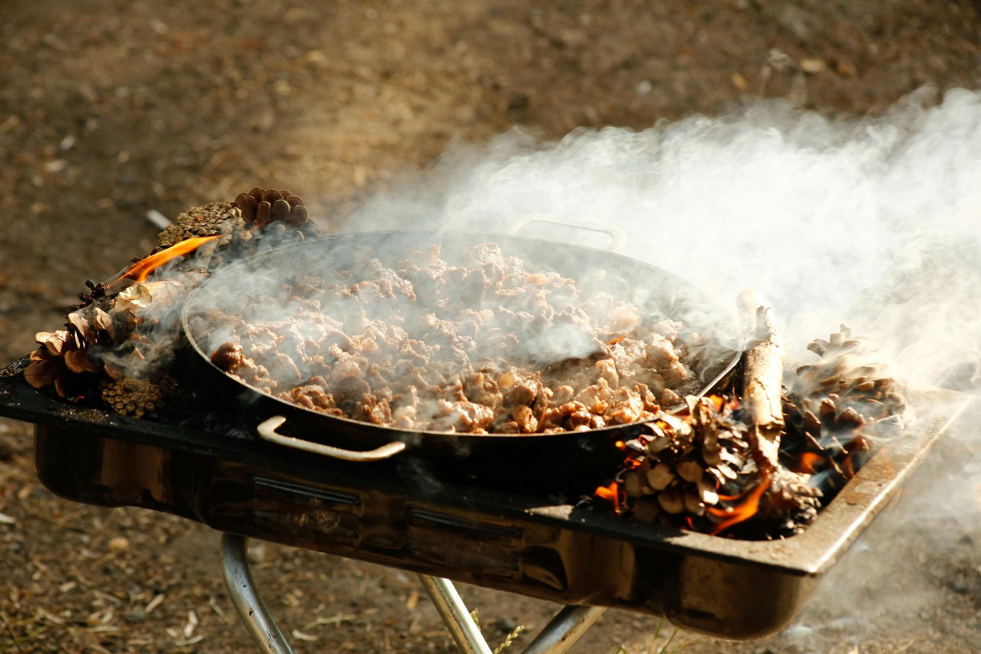Fleisch in einer Pfanne über einem rauchigen Feuer im Freien in einem Metallbehälter an einem sonnigen Tag zubereiten.