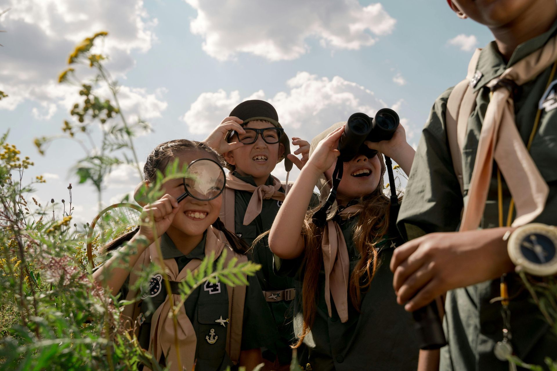 Kinder in Pfadfinderuniformen erkunden lächelnd die Natur mit Lupe und Fernglas auf einem Feld.