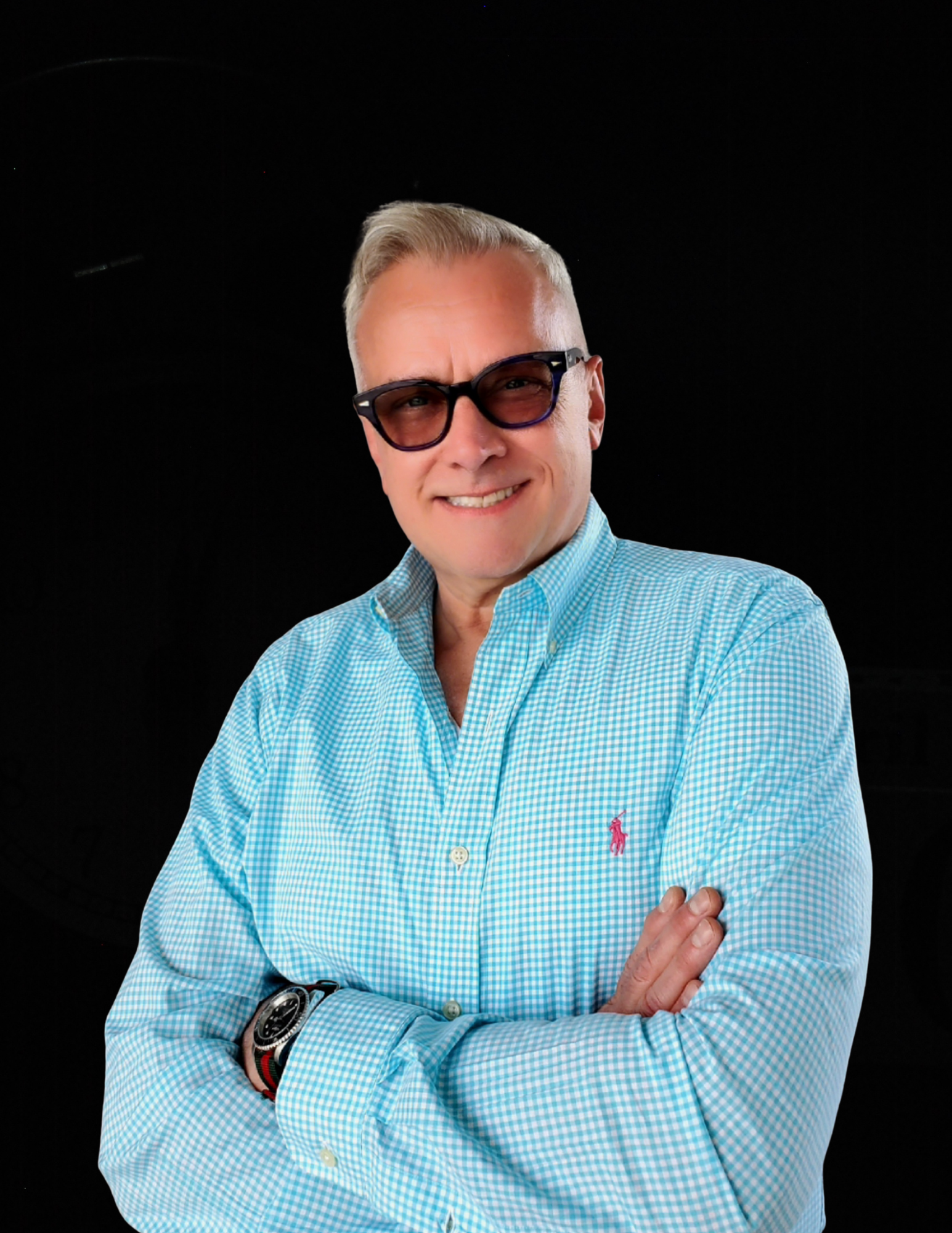 A man wearing sunglasses and a blue shirt is standing with his arms crossed.