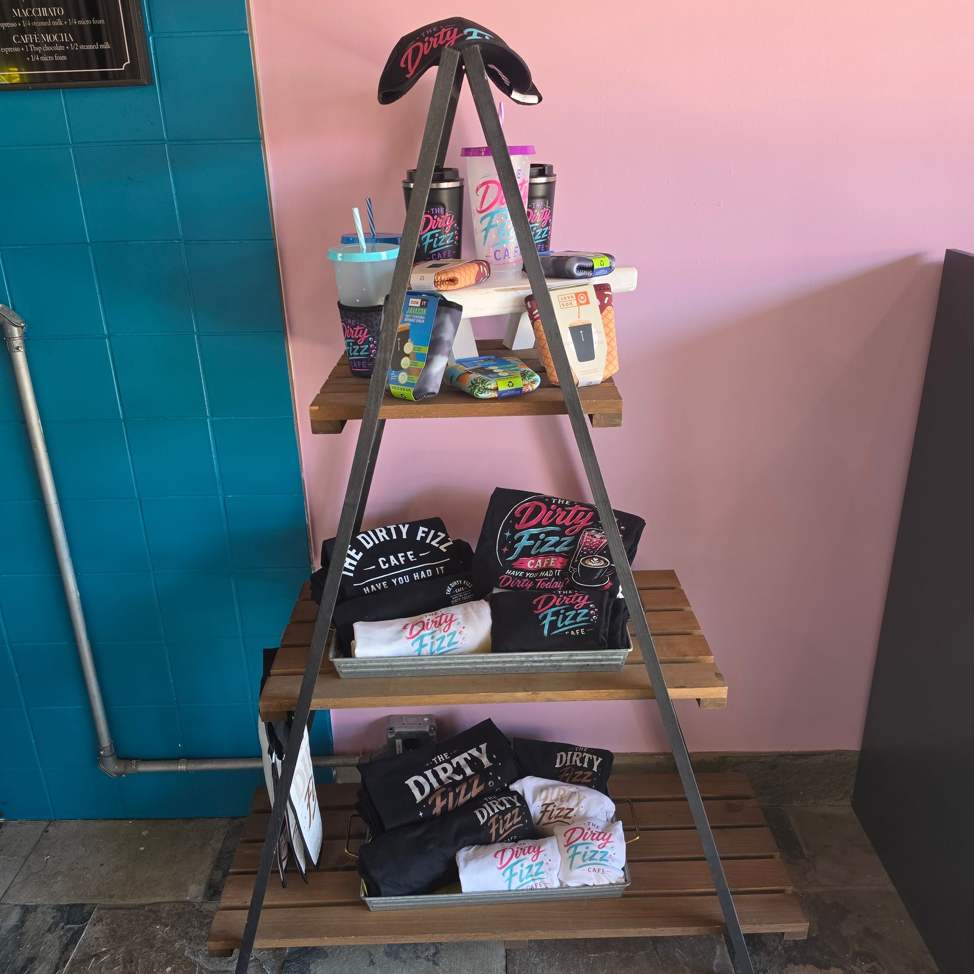 A small three-tier display shelf with black and pink T-shirts, drinks, and accessories against a pink wall.