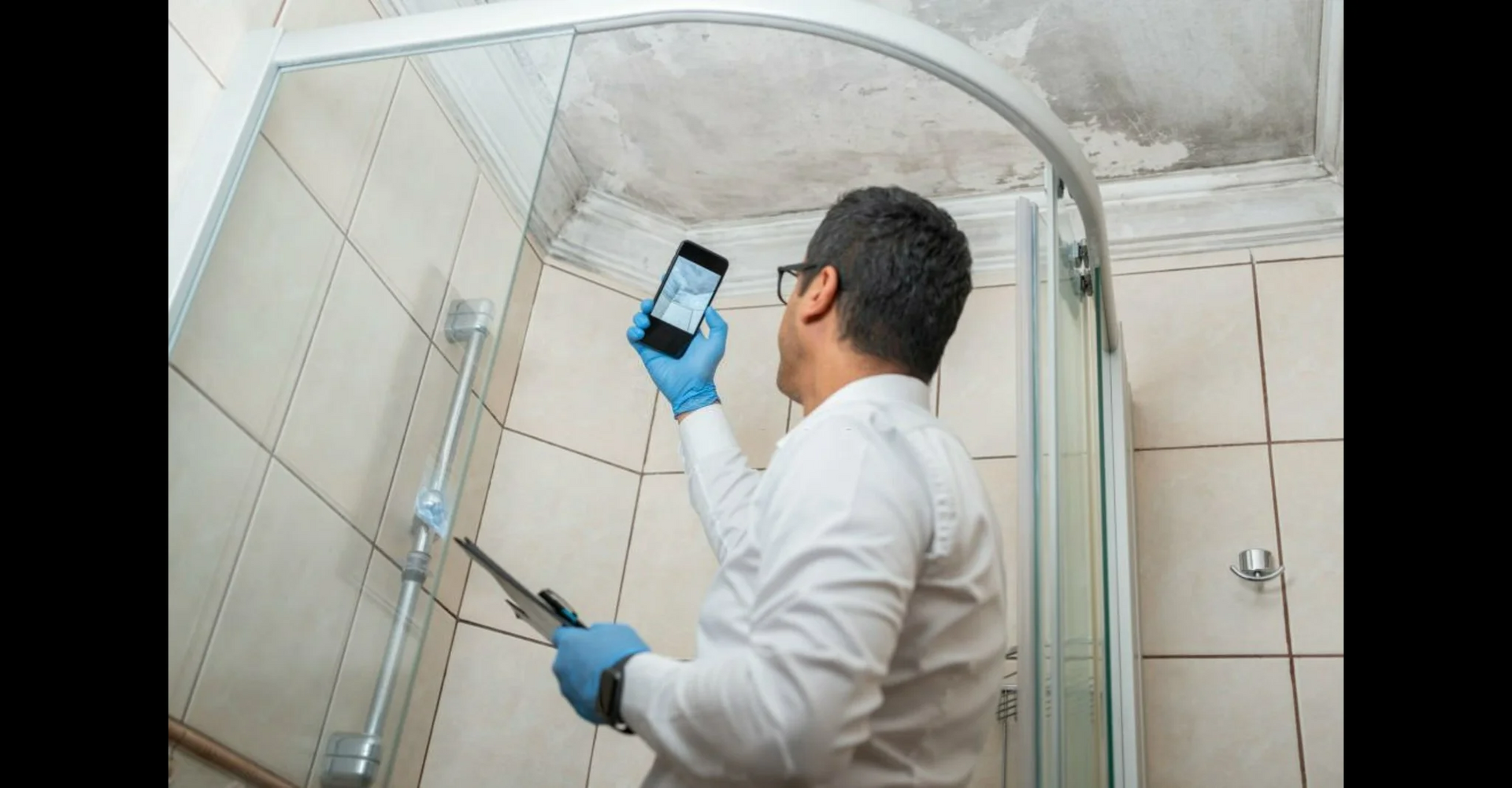A person in a white shirt and blue gloves holds a phone up to photograph water damage on a bathroom ceiling.
