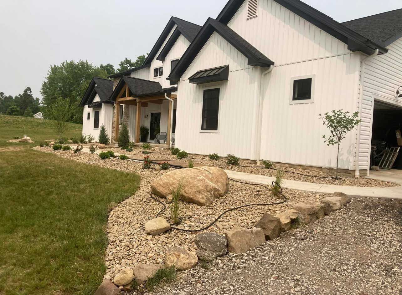 A modern white farmhouse with black trim and roof, featuring a gravel yard, stone garden borders, and a large boulder.