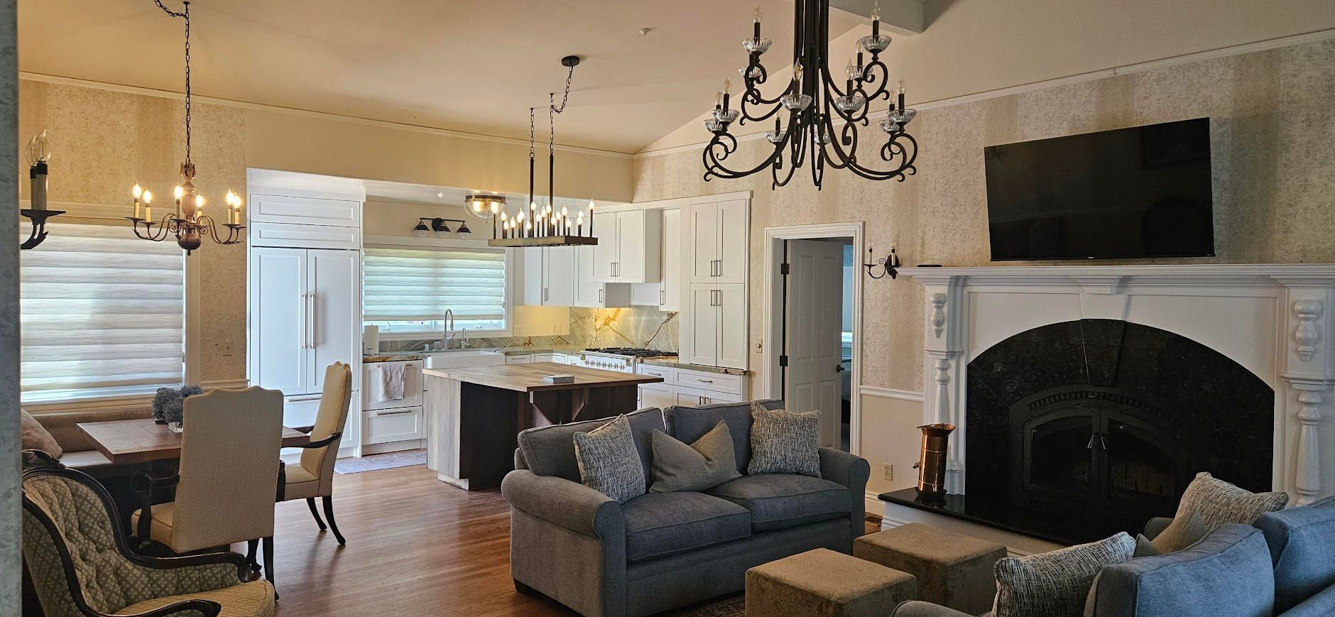 Open living room with gray sofa, fireplace, chandelier, and a view into a bright kitchen
