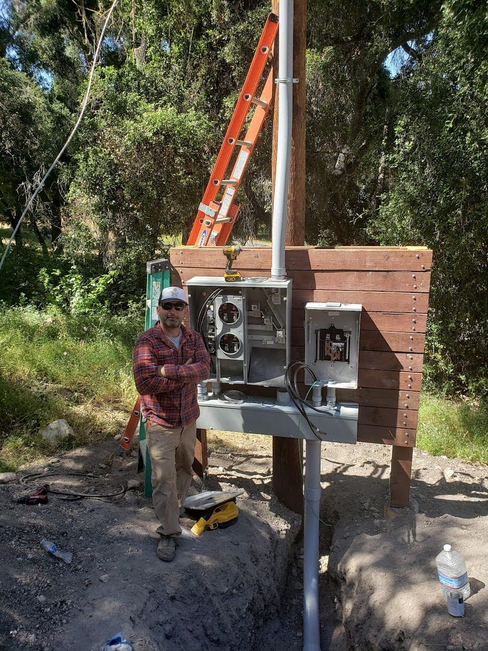 A person stands beside an electrical utility box mounted on a wooden frame outdoors, with a ladder leaning against a pole.