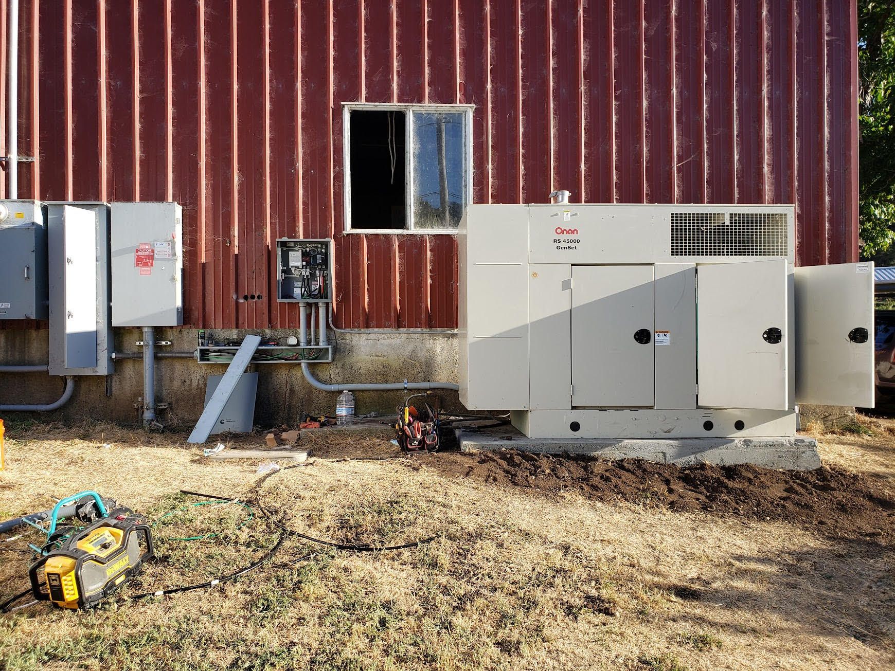 A large outdoor backup generator sits on a concrete pad next to a red metal building with electrical control boxes.