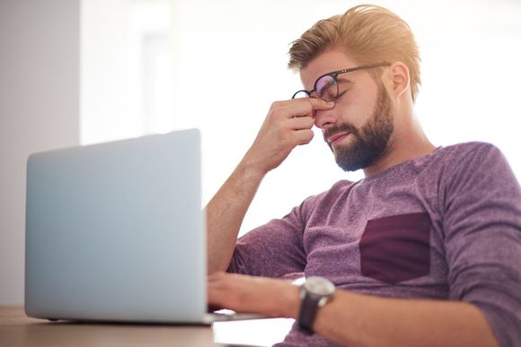 Man sitting at a laptop, rubbing his eyes in a bright room