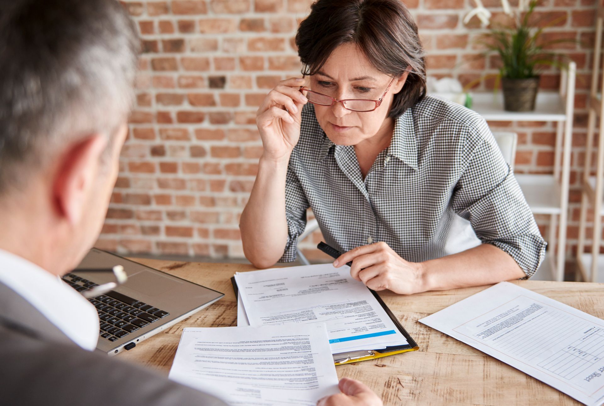 Two professionals sit at a wooden desk reviewing documents in an office with a brick wall.