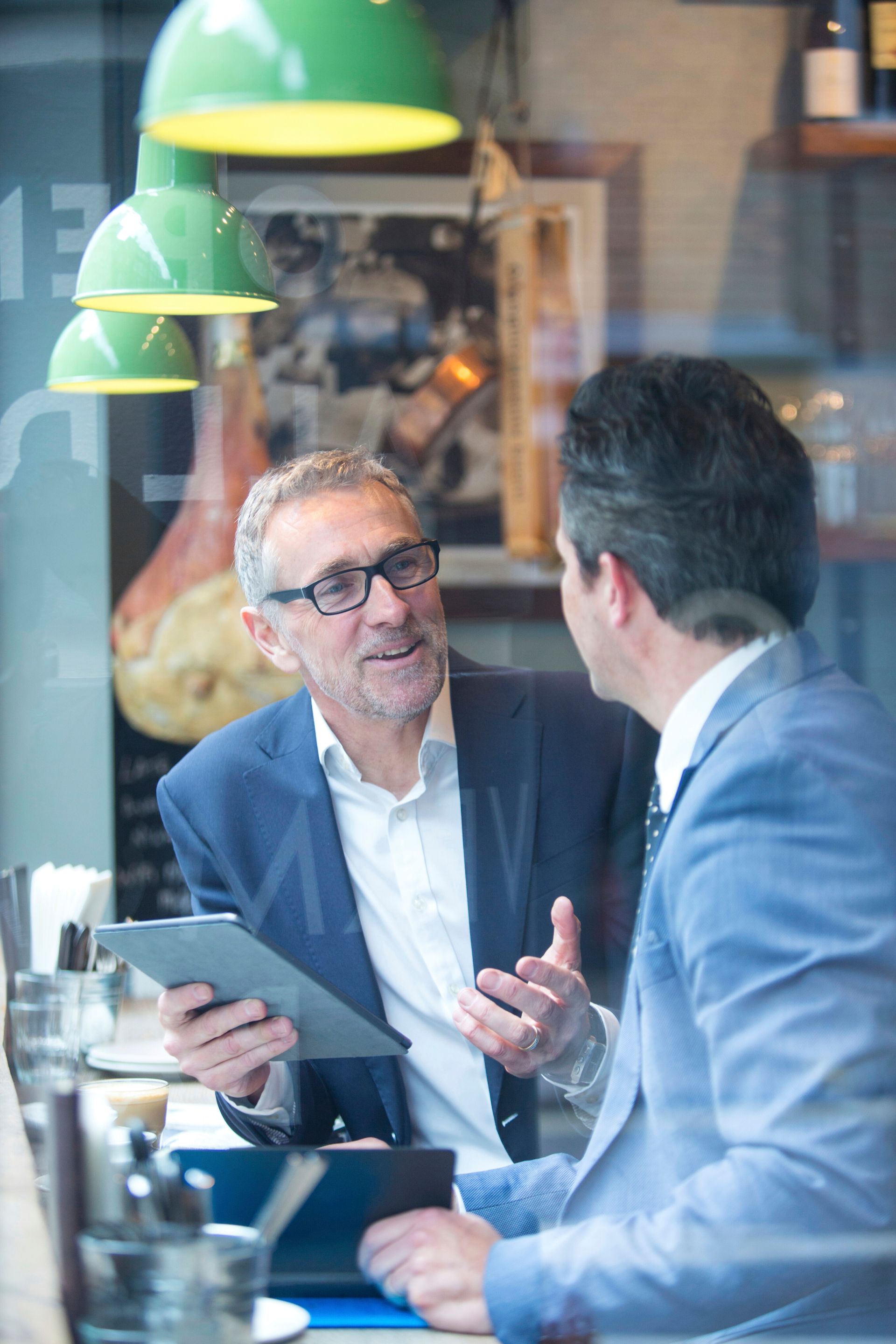 Two professionals discussing at a café table, one holding a tablet, with green hanging lamps overhead.