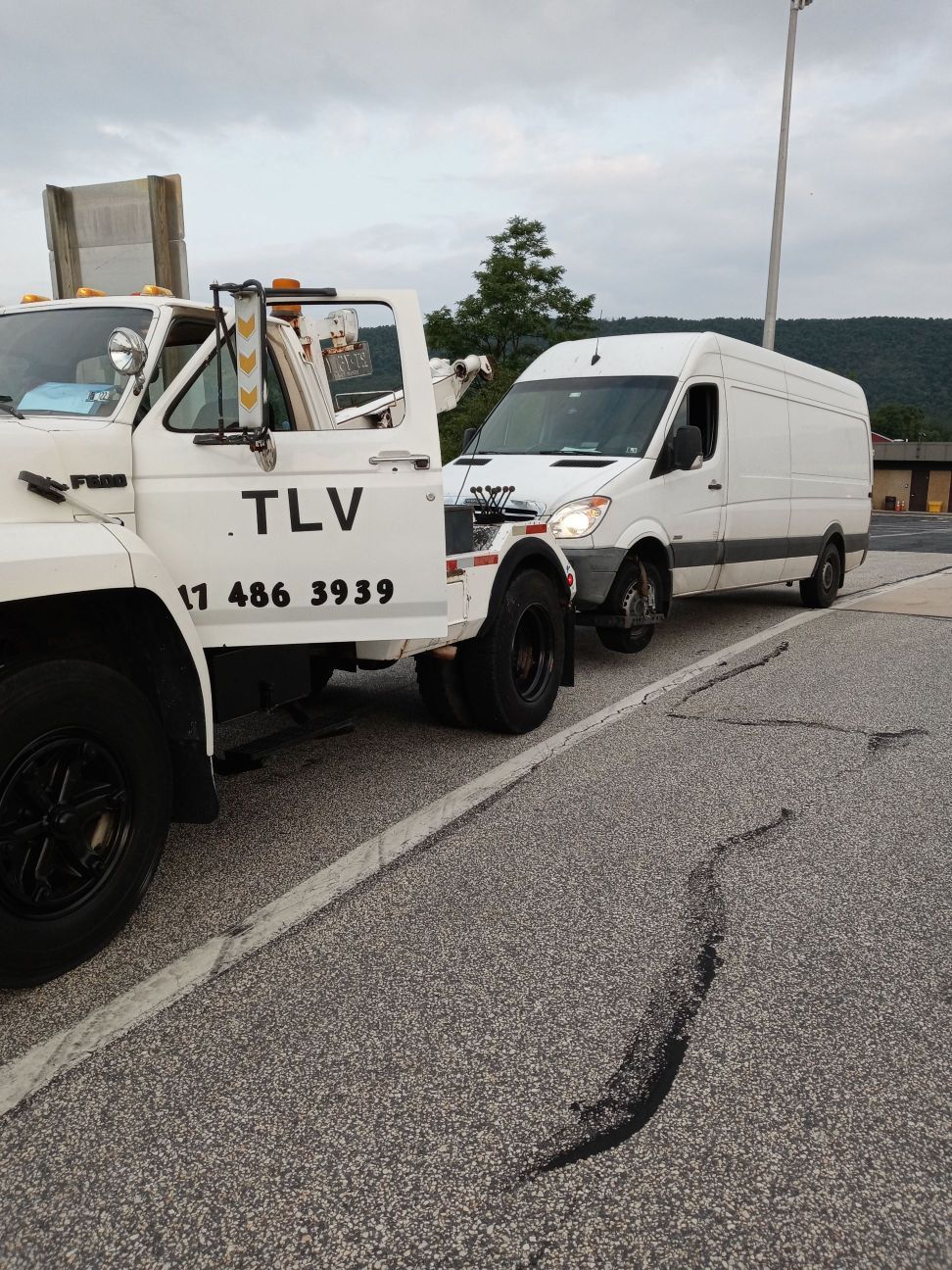 White tow truck towing a white van on a paved road. The truck has the letters 