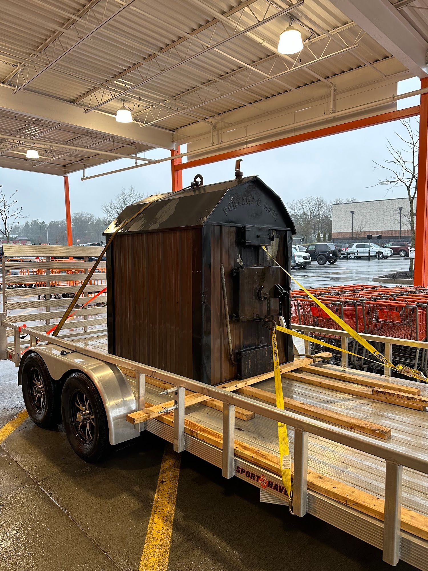 A large, dark-colored wood boiler is secured on a trailer in a Home Depot parking lot.