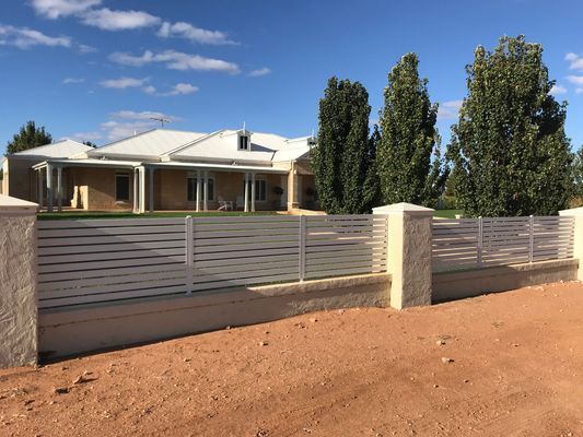 A white fence surrounds a large house with trees in the background.