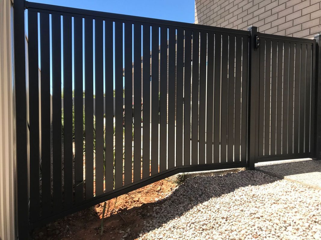 A black fence is sitting on top of a gravel driveway next to a brick building.