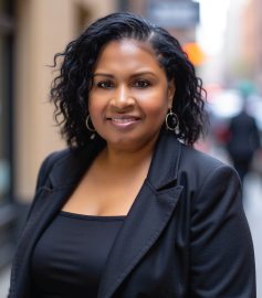 Woman in black blazer smiles, outdoors. Building in background.
