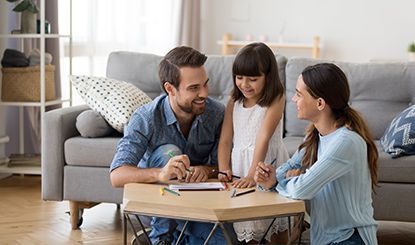 Family drawing together on a low table in a living room; smiling faces, crayons visible.