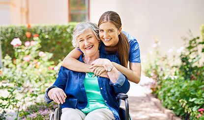 Woman embraces senior woman in a wheelchair outdoors, smiling in a garden.